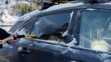 driver clears snow, ice from rear passenger window of car for clear visibility and safe driving.