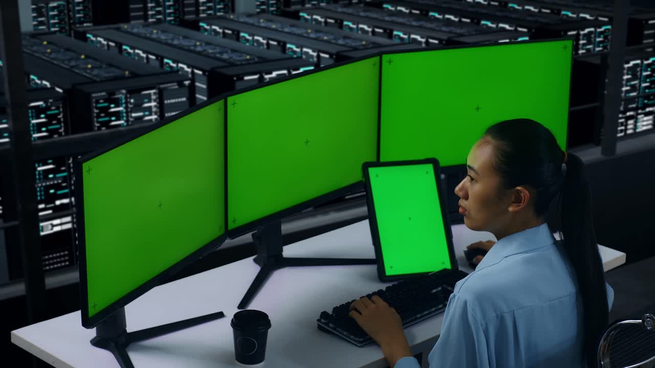 vista lateral de una mujer asiática trabajando con una tableta y varios monitores de computadora con una pantalla verde simulada en el centro de datos
