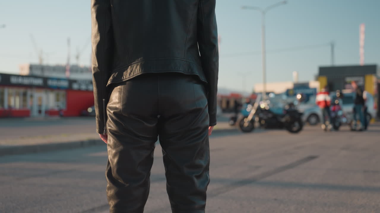 Vista posteriore di una donna in giacca di pelle che guarda un motociclista allontanarsi in lontananza, con vista sfocata di auto, edifici e persone sullo sfondo, sotto la calda luce del sole serale