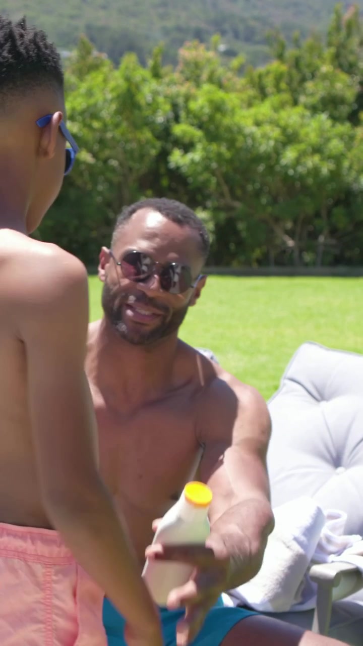 Vertical video: Leaning dad draping towel handing bottle, boy in pink trunks rubbing arms on grass
