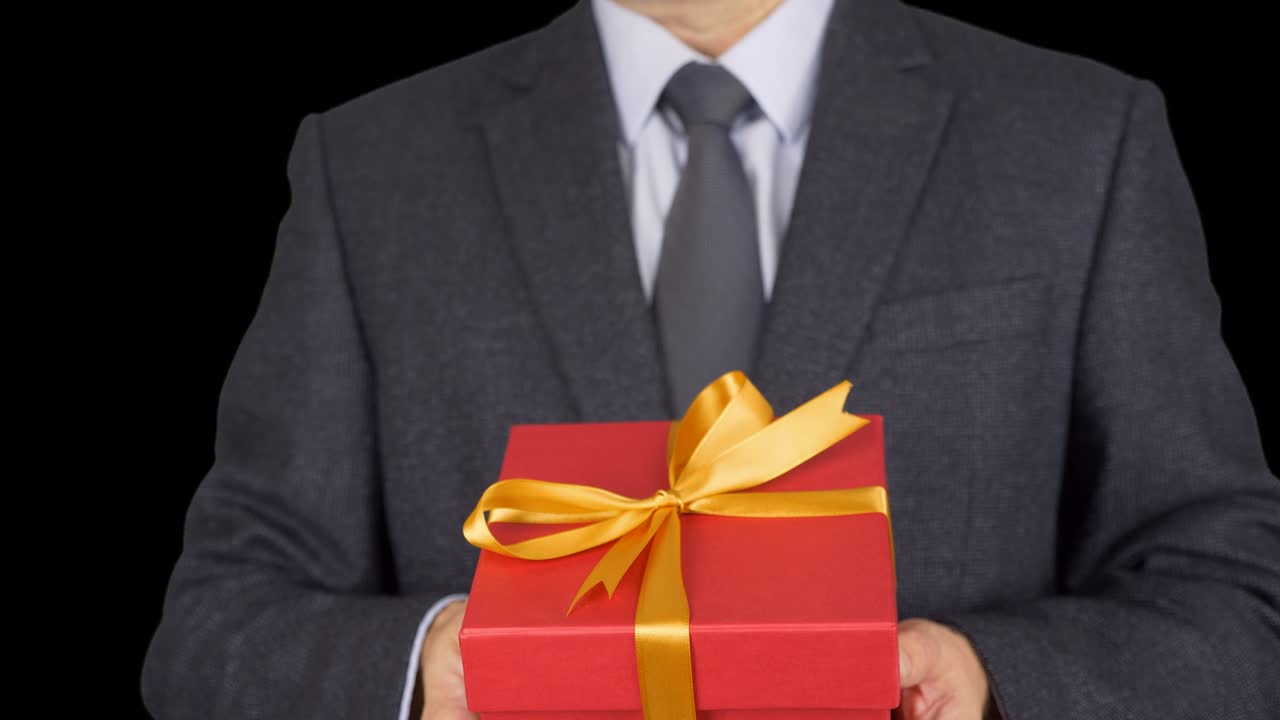 A man in a business suit with a tie is holding a red gift box. A man gives a gift. A businessman with a gift in his hands. Green screen alpha channel transparent background. From focus to unfocus.