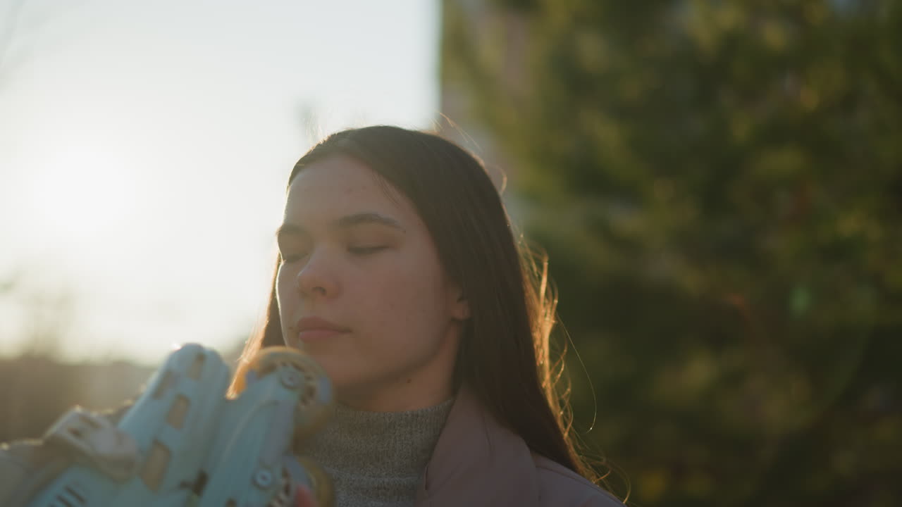 un primer plano de una mujer joven con una chaqueta de melocotón y un suéter de cuello gordo gris, de pie en un parque iluminado por el sol. ella está sosteniendo un par de patines y mirándolos con una expresión reflexiva
