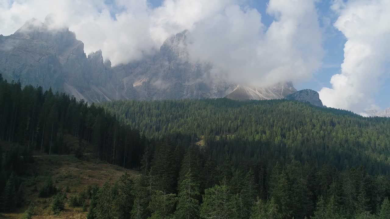 Luftfoto med drone af Dolomitternes bjergtoppe, der rejser sig over tæt fyrreskov med lave skyer, der driver hen over barske skråninger under blødt naturligt dagslys