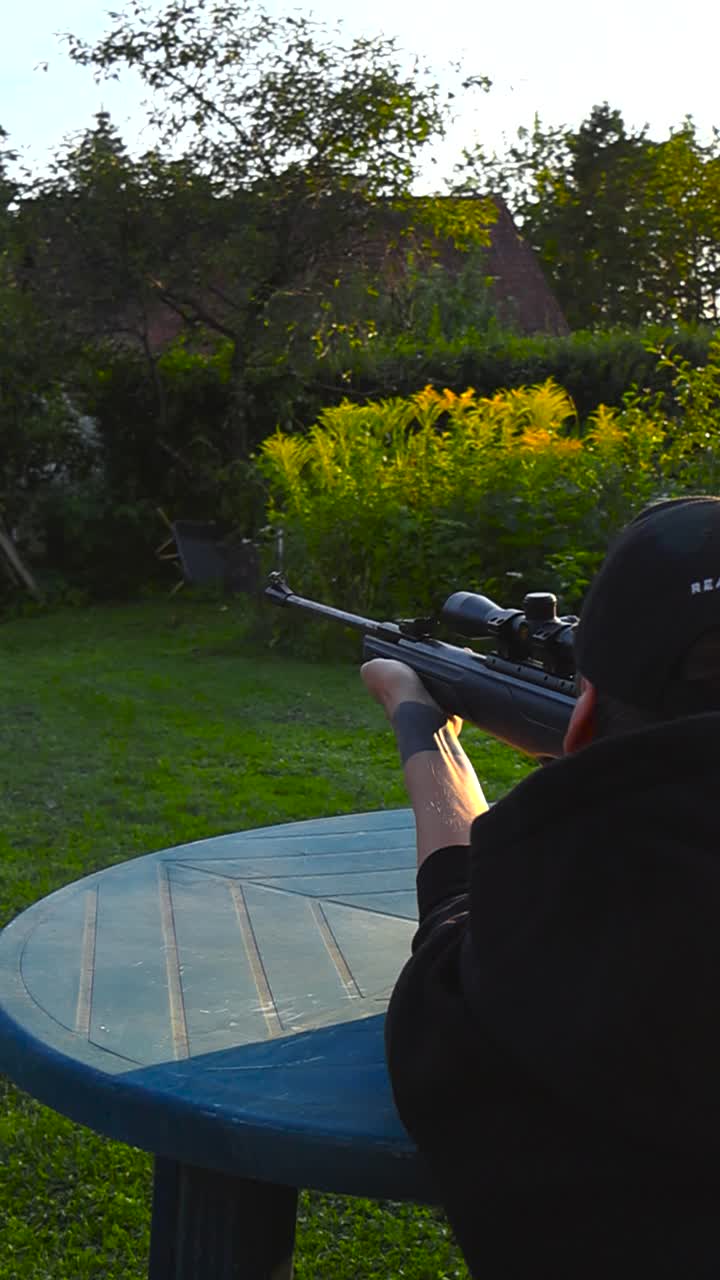 Vertical video of a white man with tattoos aiming and shooting a weapon or firearm while he squats on a table in a sunny sunset summer garden. Person is doing target practice with ammunition