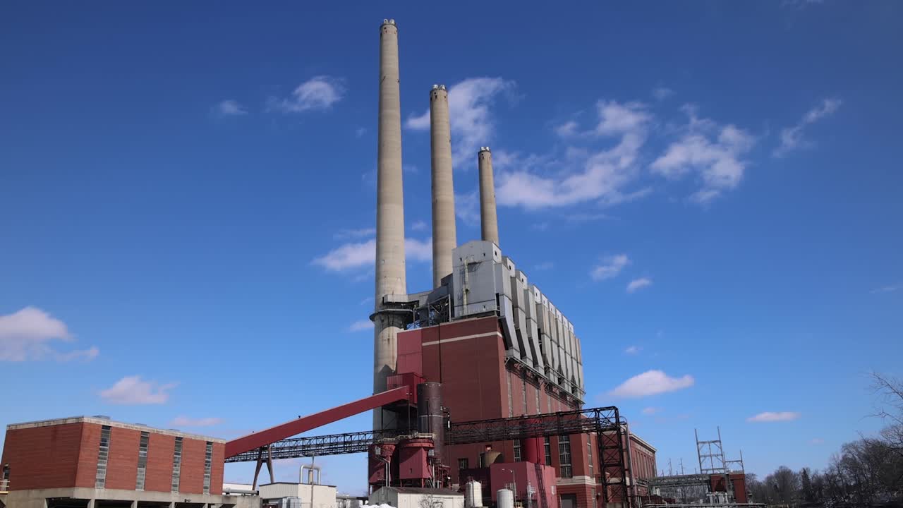 Panning shot from river dam to coal burning power plant with three smokestacks in slow motion.