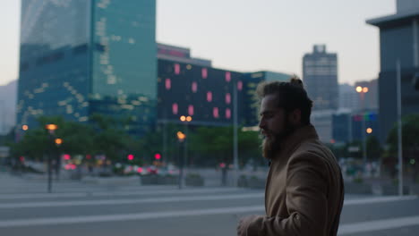 portrait of young hipster man with beard looking pensive thinking in evening urban city background waiting commuting wearing coat