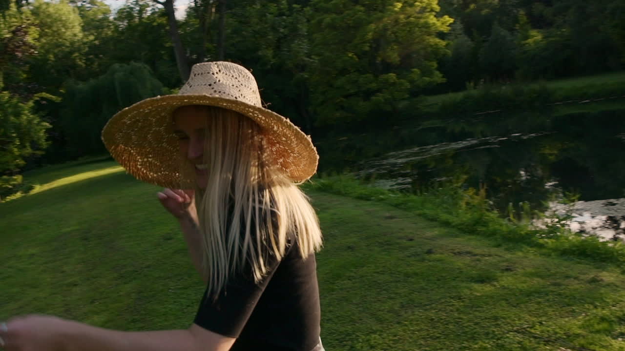 Adorable shy girl with blond hair wearing straw hat turning around, laughing and hiding from camera, summertime feeling in green garden, slow motion