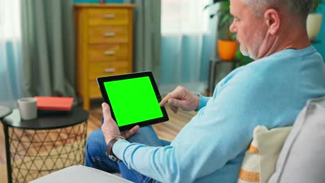 el hombre mayor en casa usa una tableta de pantalla de maqueta verde. está sentado en un sofá en su acogedora sala de estar. el hombre hace clic una vez en la pantalla y navega por el contenido.