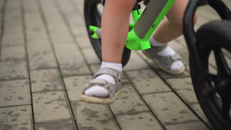 close-up of a child's legs wearing sandals with white socks while riding a bright green bicycle on a paved interlocked path