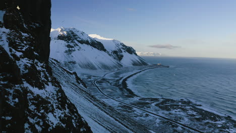 Aerial-view-backwards-over-sunlit-cliffs-with-birds,-winter-evening-in-Iceland