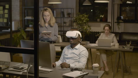 American Man Sitting At Desk In The Office Wearing Virtual Reality Glasses While A Woman Employee Is Controlling Him With Tablet