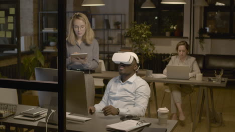 american man sitting at desk in the office wearing virtual reality glasses while a woman employee is controlling him with tablet