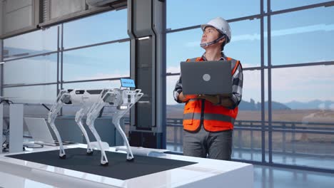 cuerpo lleno de ingeniero masculino asiático con casco de seguridad mirando una computadora portátil y mirando a su alrededor mientras está de pie en una fábrica de alta tecnología, analizando el concepto de máquina robótica