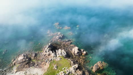 Niebla-Procedente-Del-Mar-Sobre-Un-Promontorio-Rocoso-Con-Agua-Azul-Cristalina,-Fotografía-Cenital-Brillante-Con-Niebla-Que-Se-Mueve-Lentamente