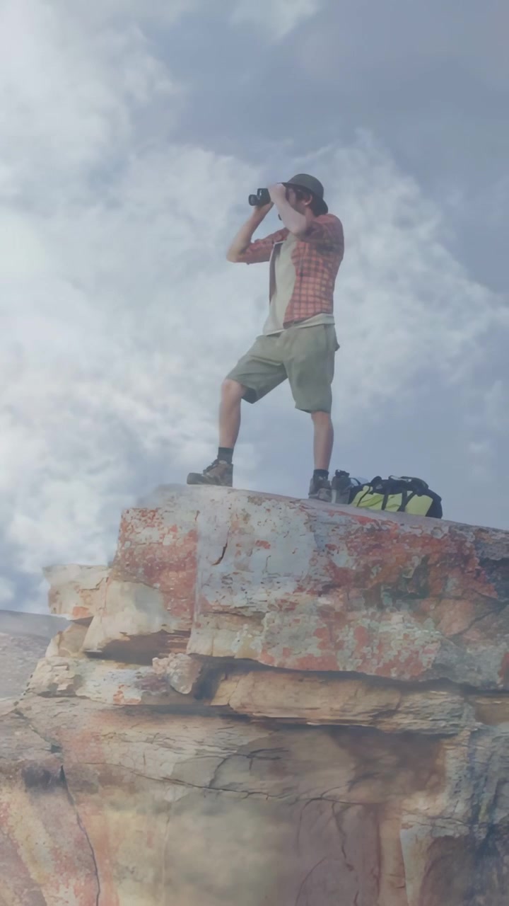 Video verticale: Uomo che alza il binocolo per cercare un sentiero da una sporgenza rocciosa, con cappello a tesa larga e zaino.