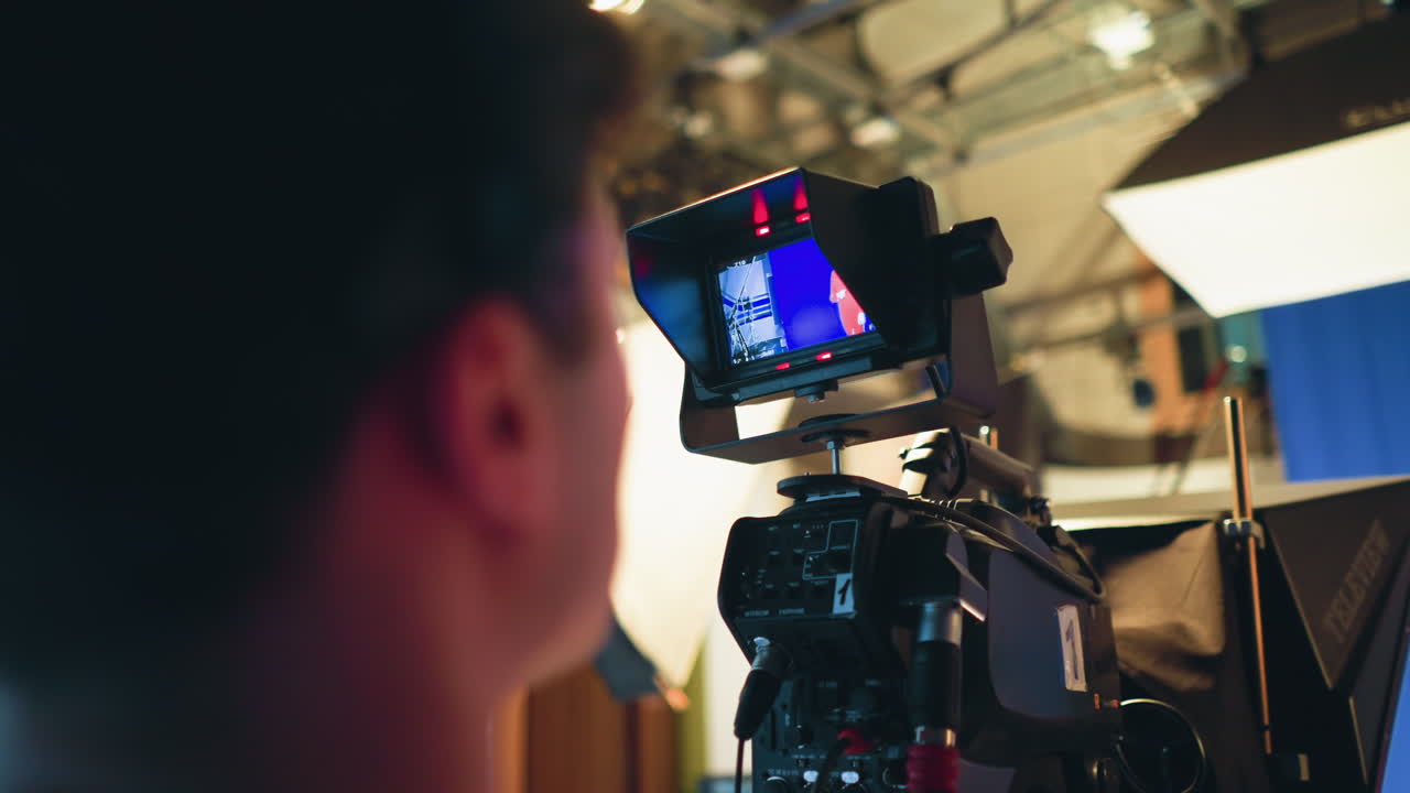 Cameraman closely monitors camera screen while filming presenter in red dress on blue screen stage during television studio shoot, surrounded by professional lighting setup and broadcast production equipment