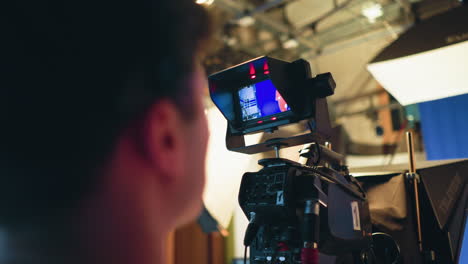cameraman closely monitors camera screen while filming presenter in red dress on blue screen stage during television studio shoot, surrounded by professional lighting setup and broadcast production equipment