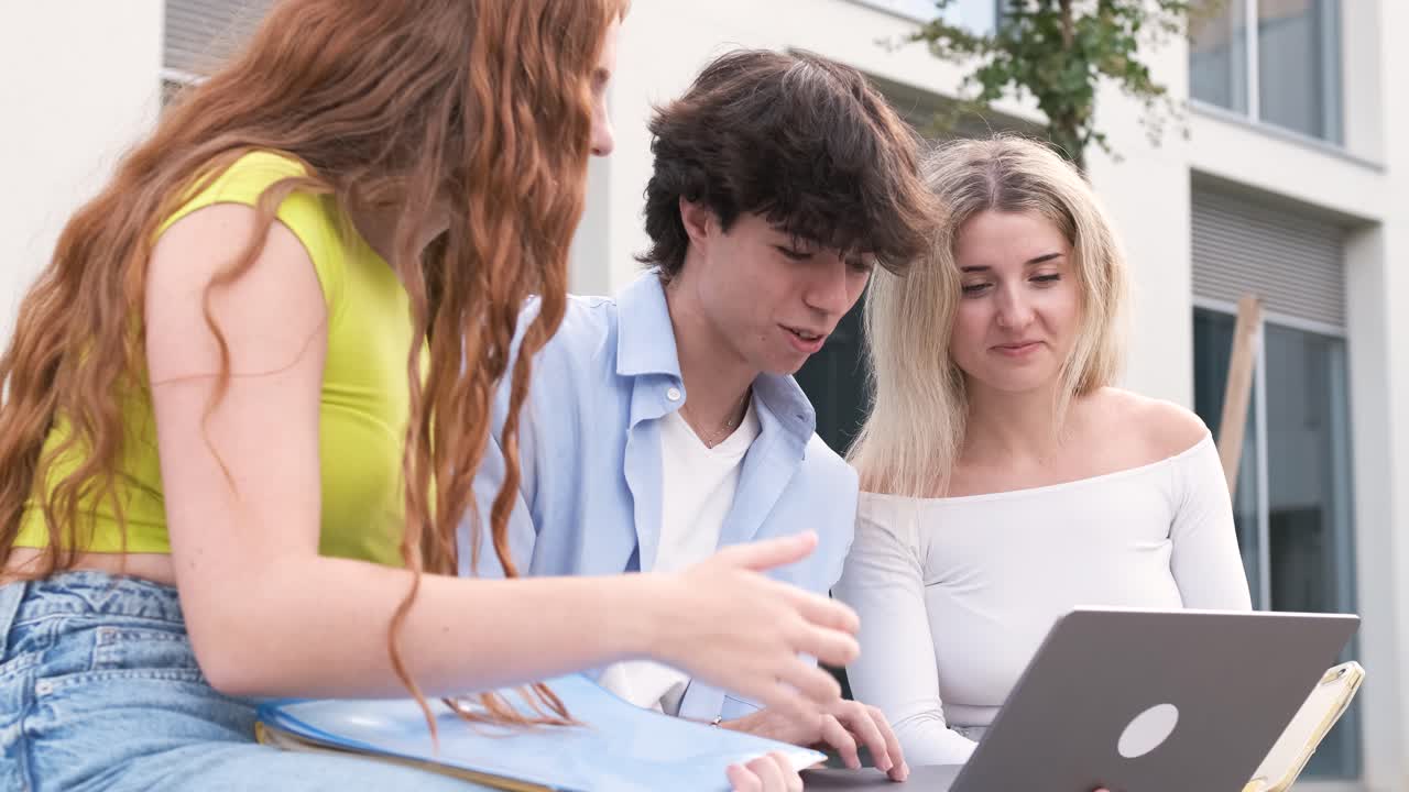 Smiling young friends using laptop outside university building