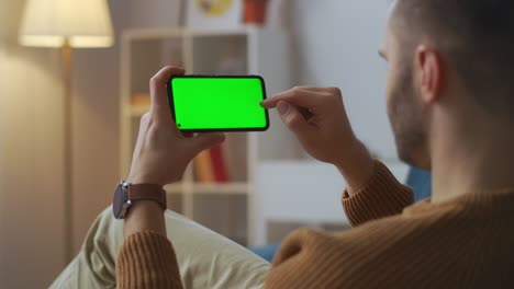 male user is tapping on screen of smartphone, holding horizontally, sitting in apartment, new app for mobile phones