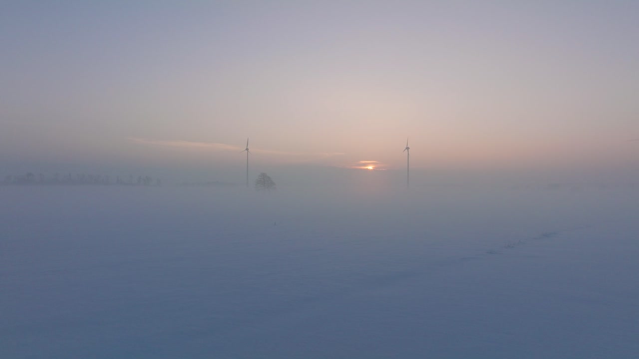 풍력 발전소에서 재생 에너지를 생성하는 풍력 터빈의 공중 전망, 안개로 가득 찬 눈으로 가득 찬 시골 풍경, 황금 시간의 빛이 있는 화창한 겨울 저녁, 넓은 드론 샷이 앞으로 낮게 움직입니다.