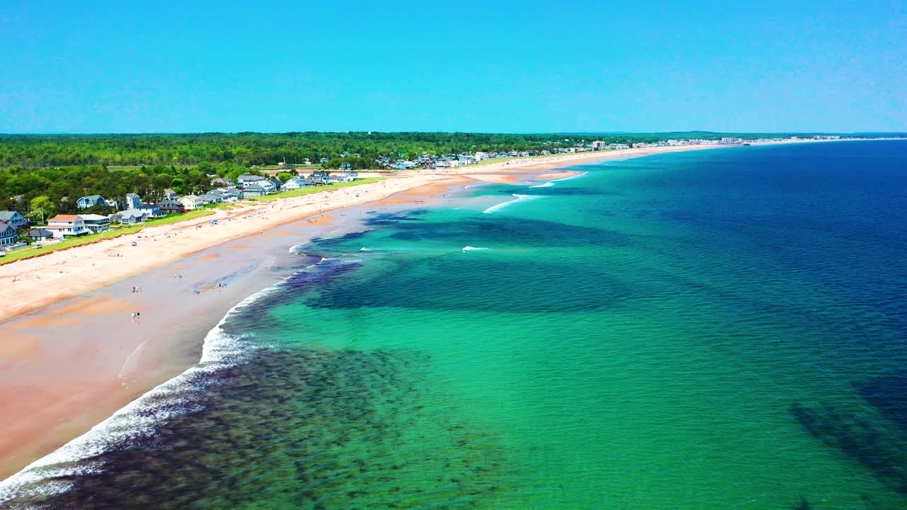 Drone flying above Saco, Maine coastline shows people enjoying peak-season beach life. Sand full of tourists, vacation homes nearby, waves crash on shore. A beautiful summer day on the Atlantic.
