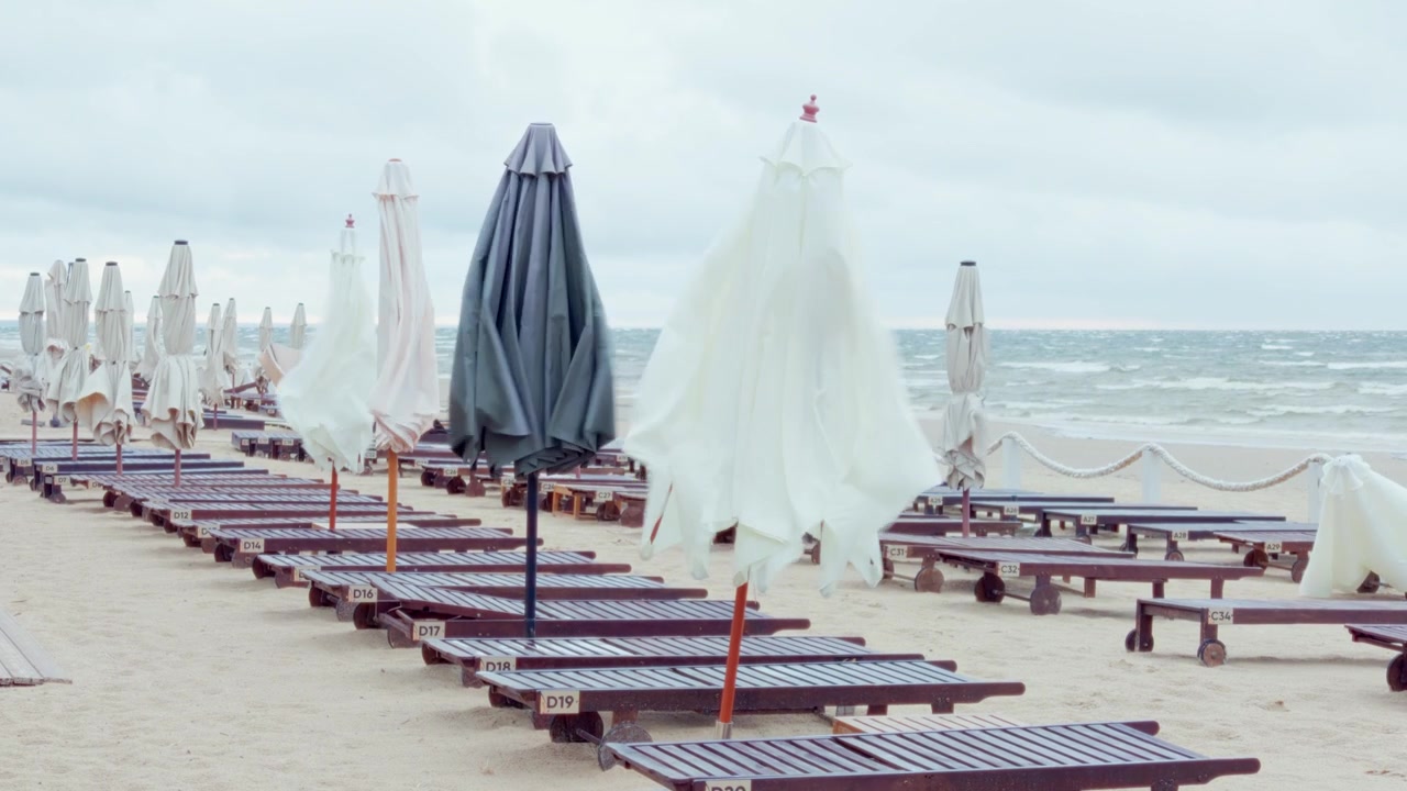 Una ripresa statica mostra file di sedie a sdraio in legno vuote e ombrelloni piegati sulla costa sabbiosa del Baltico durante una giornata estiva ventosa e nuvolosa nella località di Jurmala
