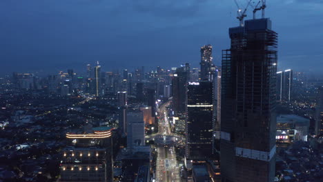 Wide-rising-pedestal-aerial-shot-of-Jakarta-city-cityscape-at-night-with-tall-skyscrapers-and-busy-traffic-around-Selamat-Datang-monument-roundabout