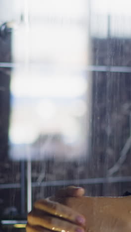 young woman with blonde hair bun washes shoulder and body under hot running water in shower cabin against marble tile daily routine slow motion