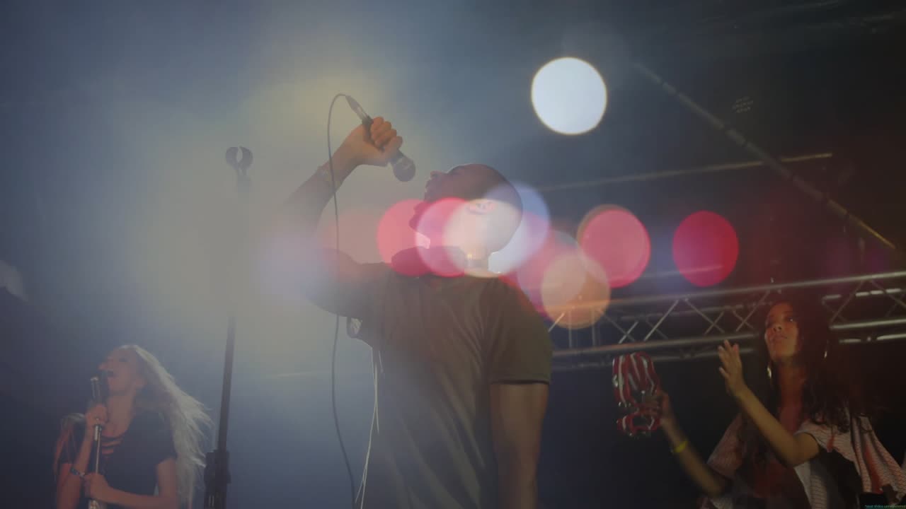 Cantante principal avanzando al centro del escenario y agarrando el micro para el estribillo mientras luces de colores se mueven por encima.