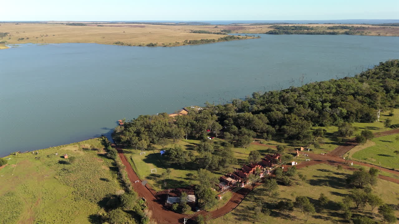 Drone shot captures riverside settlement along the Paraná River between Misiones and Corrientes, Argentina, with forested shore and vast rural landscape under clear sky, real time, aerial view