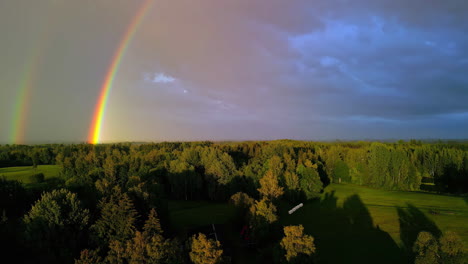 aerial drone tilt up shot of two rainbows falling into mystic forest along with rural cottages after rainfall