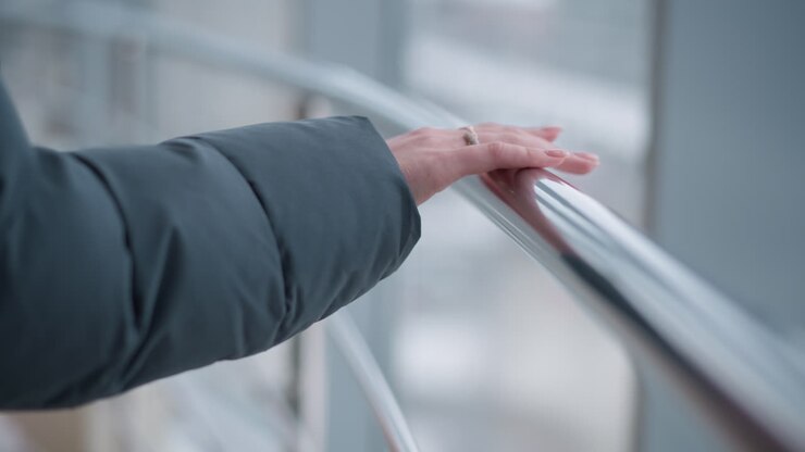 Primo piano della mano di una signora adornata con un anello, posata sul corrimano di una scala mobile in discesa, con uno sfondo di vetro che mostra un paesaggio esterno in un luminoso ambiente urbano.
