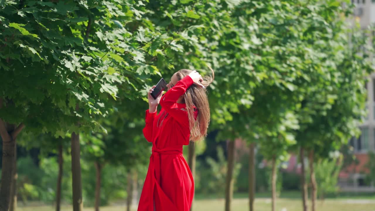 Young woman in beautiful red dress. Lovely female talking on the phone on green trees background. Attractive lady outdoors. Slow motion.
