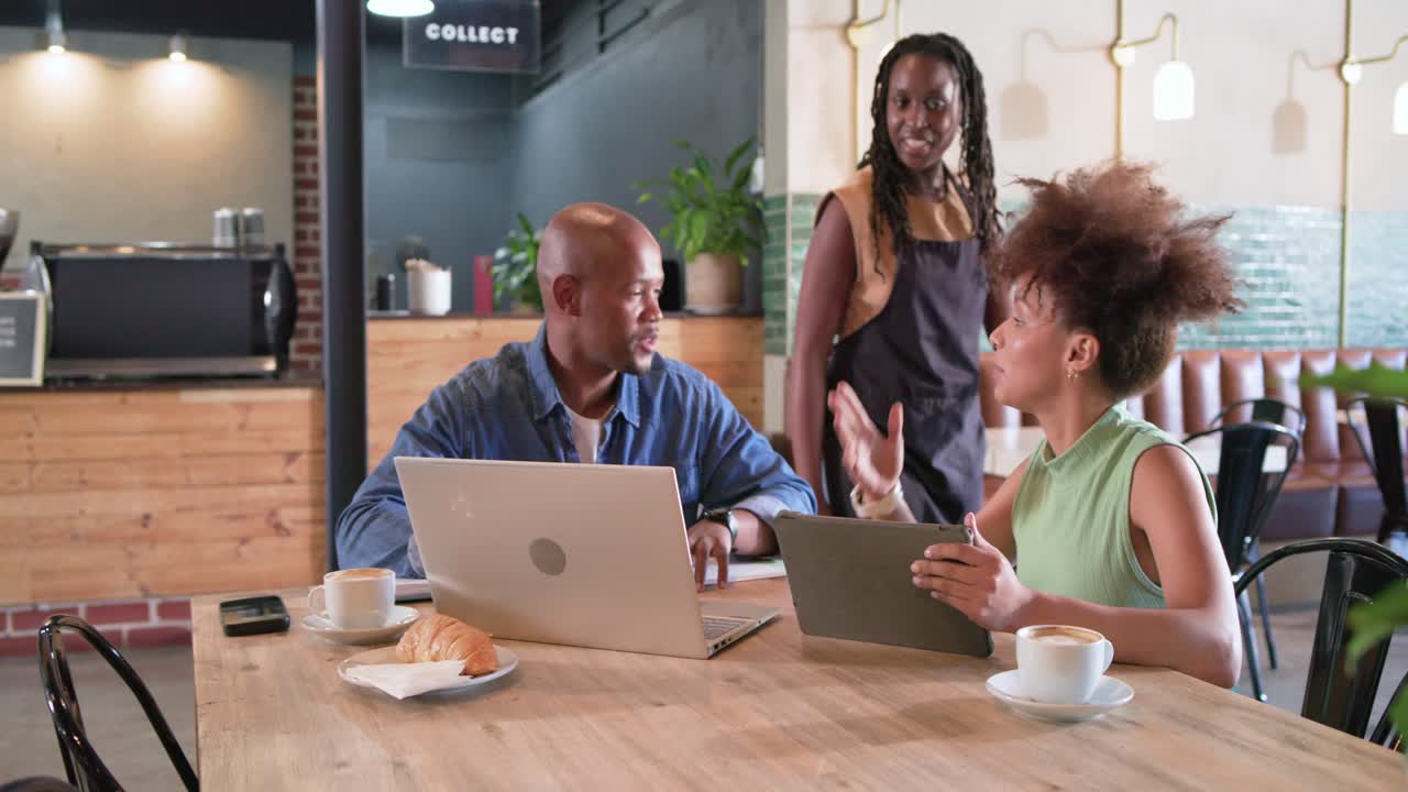 African American pair at cafe using devices, turning as barista approaching to chat, then returning