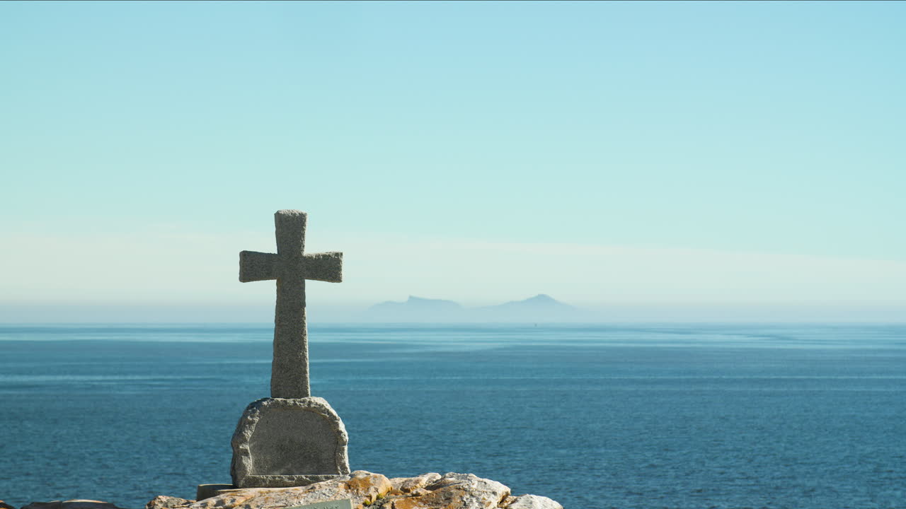 Tombstone memorial in left of frame overlooking calm ocean water with heatwaves visible and layer of mist and blue mountains in distant horizon on clear sunny day
