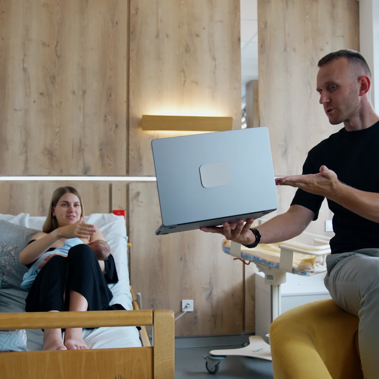 Caucasian couple having a newborn in the maternity hospital. Woman breastfeeding a baby and husband shows her something on laptop