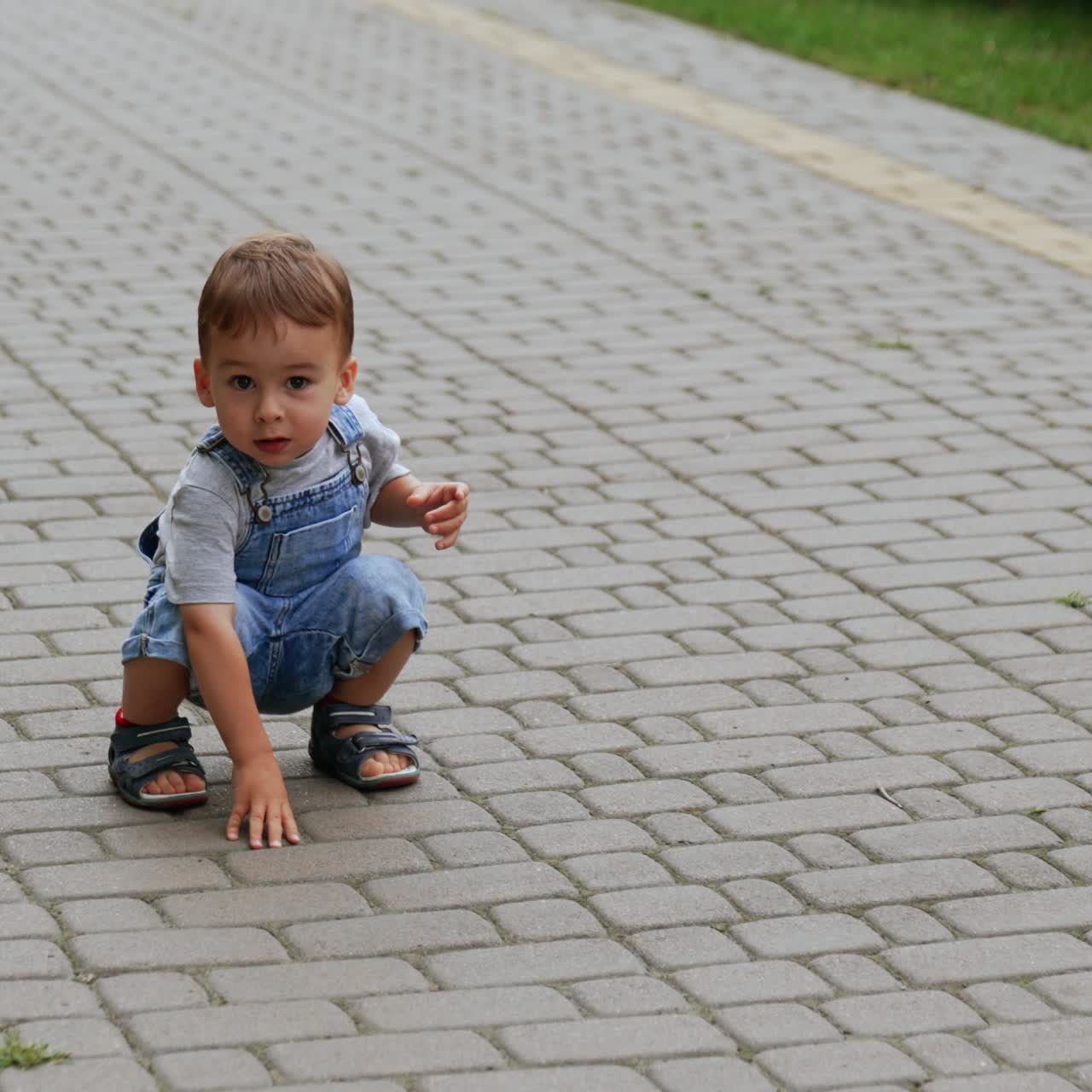 Cute baby boy in jeans romper squats on the paved road. Interested kid touches the tiles on the ground