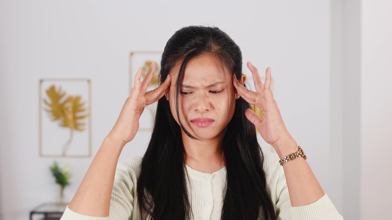 A woman appears confused and worried, holding her head in a well-lit room with minimal decor