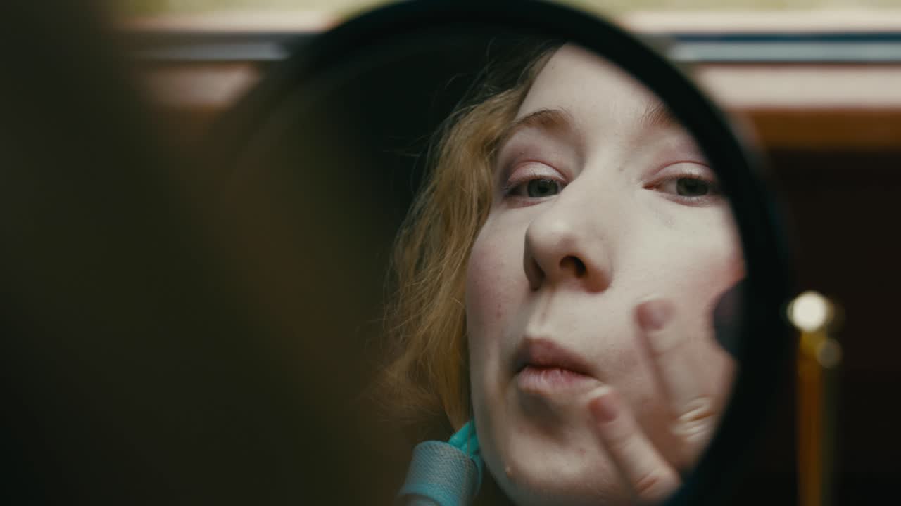 Young blonde girl applying powder makeup with brush while looking into mirror, close up