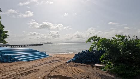 wide shots of workers resting near stacked blue pipes on a sandy beach, shaded by trees, with bright daylight and a calm sea backdrop