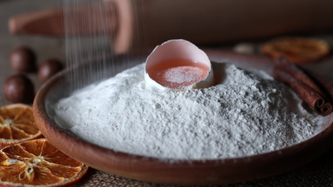 White flour dust fall to egg yolk inside half of egg shell atop flour pile in plate