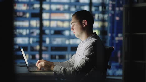 night work a man sits at a table and prints on a laptop against the background of the night city