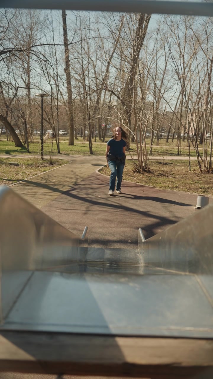 Close up of woman interacting with playground equipment sliding down and walking away in outdoor park setting. Her hand rests on metal slide while her hair moves freely capturing natural
