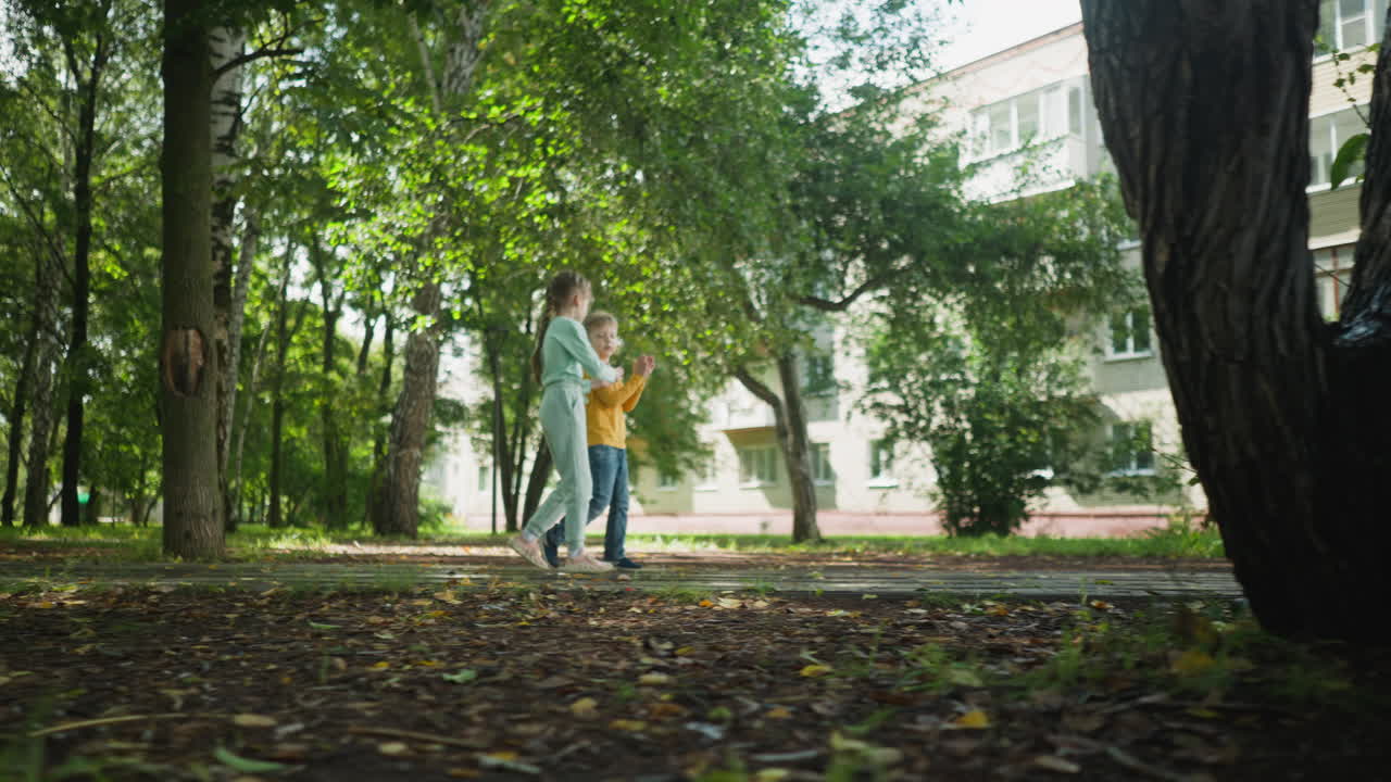 Young boy and girl walking together on park path near residential apartment building, surrounded by trees and nature, enjoying relaxed moment while talking and interacting in peaceful neighborhood