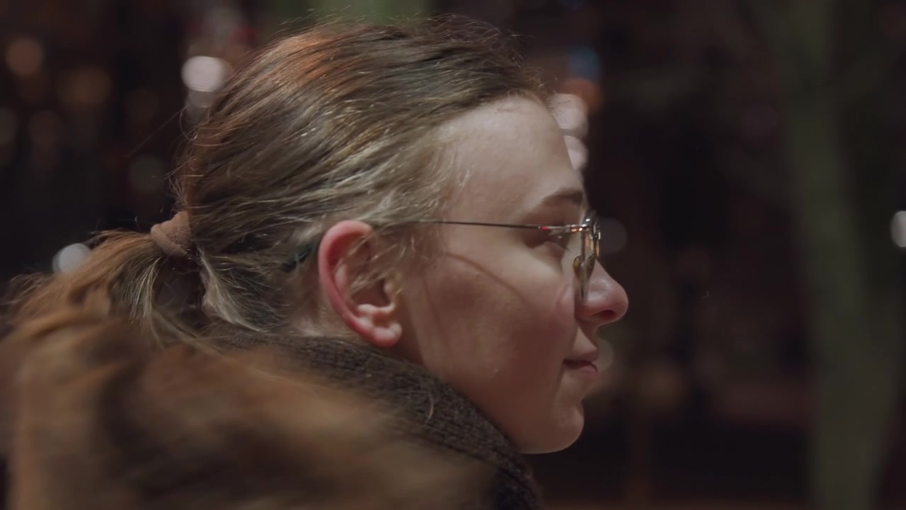 Night park closeup of white woman gently gathering hair into ponytail, snowflakes caught in strands, glasses framed face, wool scarf and coat, soft bokeh, introspective mood, subtle movement, cold