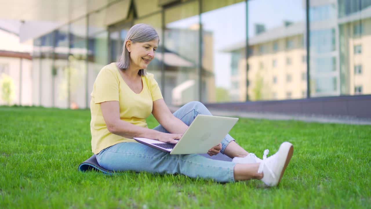 Happy senior modern woman working with laptop remotely outdoors. sitting on a grass lawn in an urban park in nature. Mature female uses a laptop on a city street