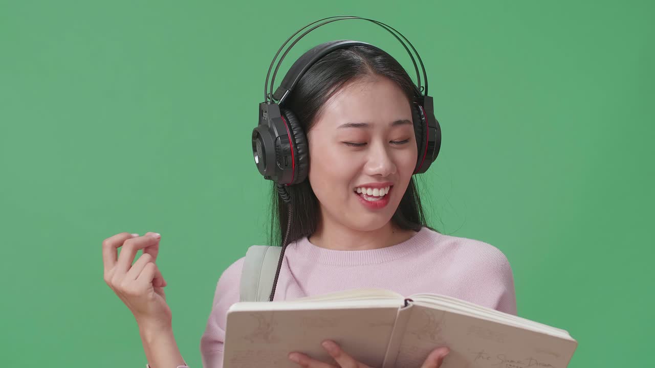 아시아 여성 학생이 책을 들고 헤드폰으로 음악을 듣고 녹색 스크린 스튜디오에서 춤을 추고 있습니다.