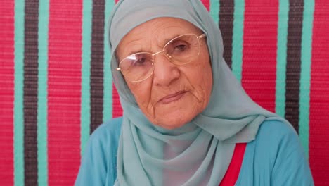elderly woman in a blue hijab sits calmly against a vibrant red and teal striped background