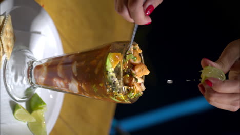 una foto en cámara lenta vertical de una mujer con las manos apretando una lima en un cóctel de mariscos servido en un vaso de la vieja moda en un restaurante tradicional en méxico