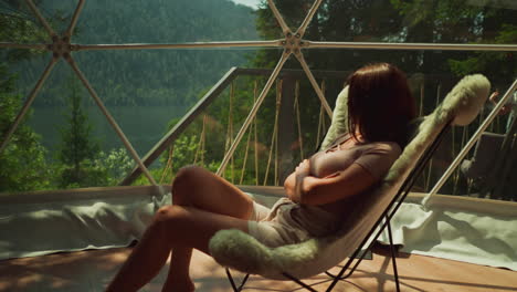 young lady in casual clothes sits on soft armchair in sunlight. woman relaxes on transparent balcony surrounded by exotic trees. picturesque mountain view out of window